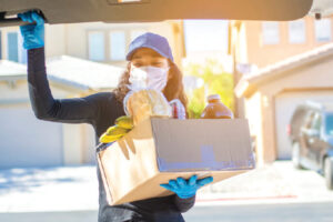 Woman wearing a mask puts a box of groceries into a car trunk